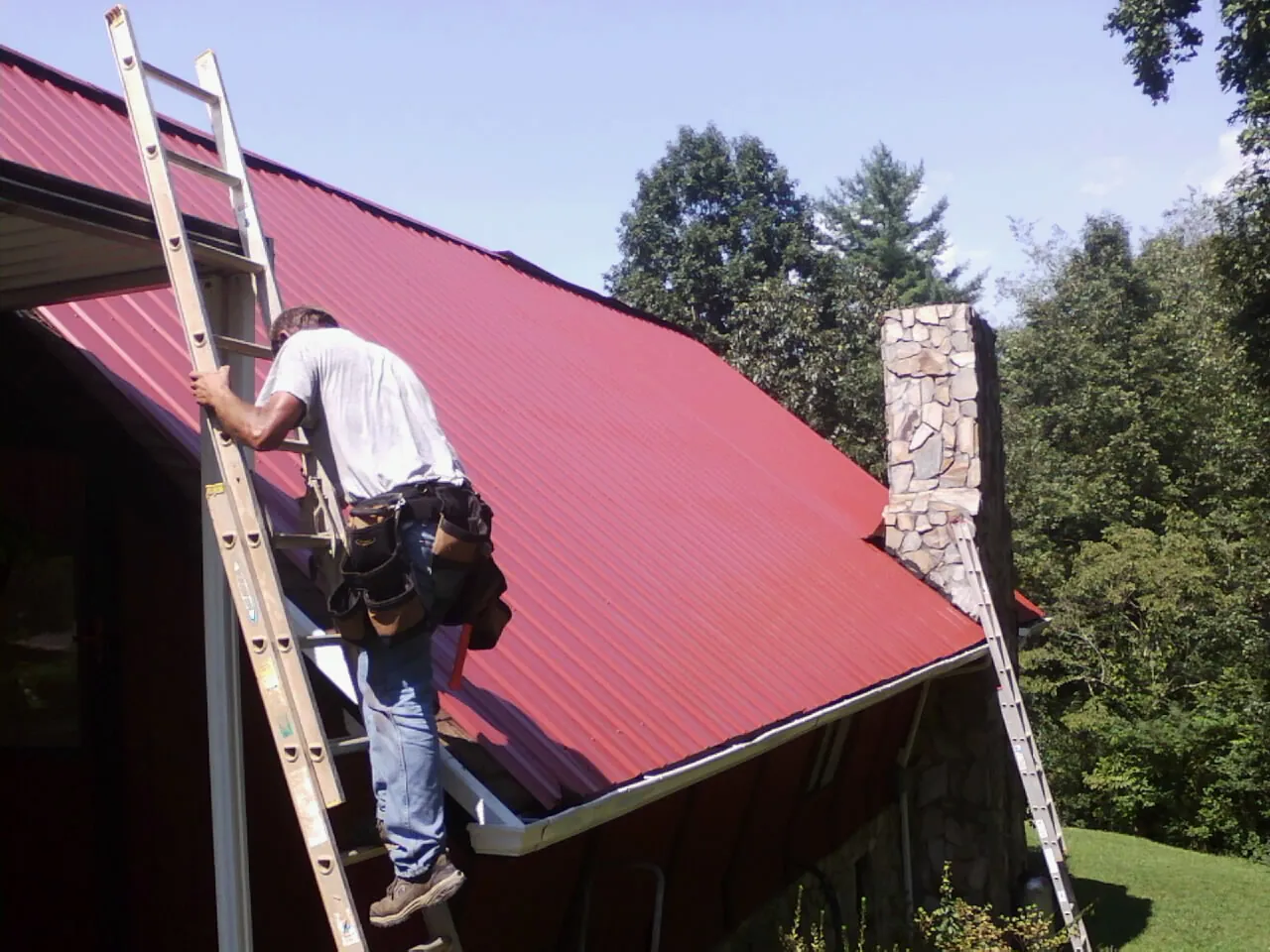 Roofer on a ladder beside a red metal roof and stone chimney — Soffit & Fascia Repair by Roofing Brothers in Berkey